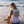 A woman sits on a sandy beach at the water's edge, viewed from behind. She is wearing a white sleeveless tank top with a large circular red and dark red graphic on the back that reads 'NAUTILIFE BEACH SEX EXTRA SAND'.