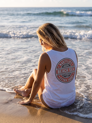 A woman sits on a sandy beach at the water's edge, viewed from behind. She is wearing a white sleeveless tank top with a large circular red and dark red graphic on the back that reads 'NAUTILIFE BEACH SEX EXTRA SAND'.