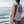 A man standing on a cliff overlooking the ocean, seen from behind, wearing a white sleeveless tank top with a large circular 'Nautica Beach Sex' graphic on the back and colorful tropical print board shorts, while holding a white surfboard.