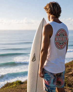 A man standing on a cliff overlooking the ocean, seen from behind, wearing a white sleeveless tank top with a large circular 'Nautica Beach Sex' graphic on the back and colorful tropical print board shorts, while holding a white surfboard.