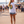 A woman stands on a sunny beach boardwalk wearing a white long-sleeve crewneck shirt with a large circular multicolor graphic on the back that reads 'OILWEAR'.