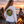 A woman standing on a beach at sunset wearing a white long-sleeve shirt with a large circular tropical wave graphic on the back that reads 'NAUULIFE'. Two other women and a campfire are visible in the background.