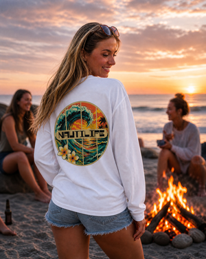A woman standing on a beach at sunset wearing a white long-sleeve shirt with a large circular tropical wave graphic on the back that reads 'NAUULIFE'. Two other women and a campfire are visible in the background.