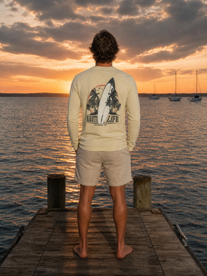 A man stands on a wooden dock at sunset facing a lake, wearing a cream long-sleeve shirt with a large surfboard and trees graphic on the back that reads 'NAIL / LIFE', paired with light beige shorts.