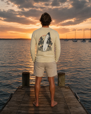 A man stands on a wooden dock at sunset facing a lake, wearing a cream long-sleeve shirt with a large surfboard and trees graphic on the back that reads 'NAIL / LIFE', paired with light beige shorts.